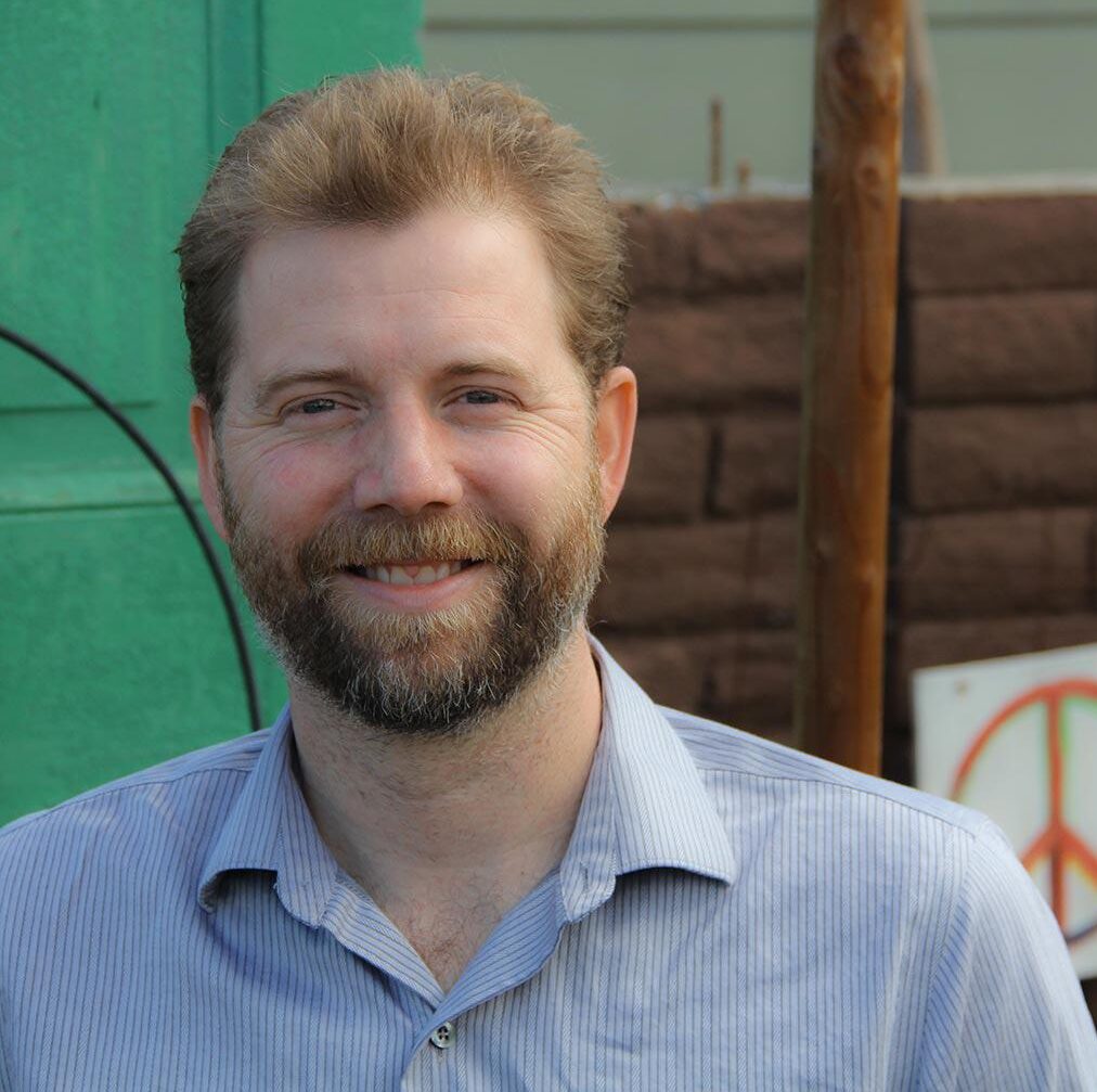 Photo of the Chief Operating Officer at Get Community Ryan Canton who has a beard is wearing a blue dress shirt and is standing in front of a green door brown bricks and an orange peace sign
