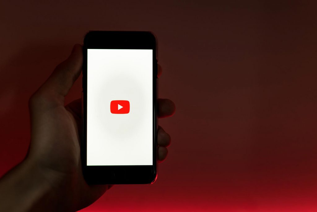 black persons hand in front of a black and red background holding a smartphone with the white and red YouTube loading image on the screen